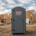A portable toilet stands in front of a wooden building under construction at a worksite, with construction materials scattered around.